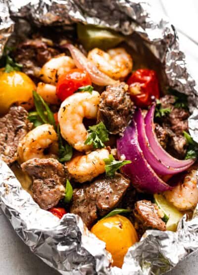 close up shot of a foil pack filled with steak bites, shrimp, red onions, and colored grape tomatoes.