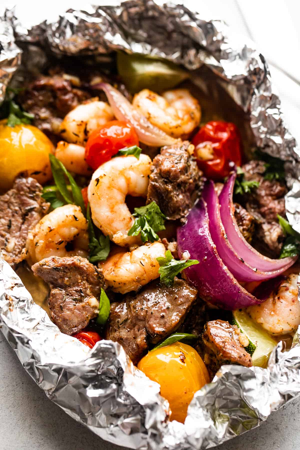 close up shot of a foil pack filled with steak bites, shrimp, red onions, and colored grape tomatoes.