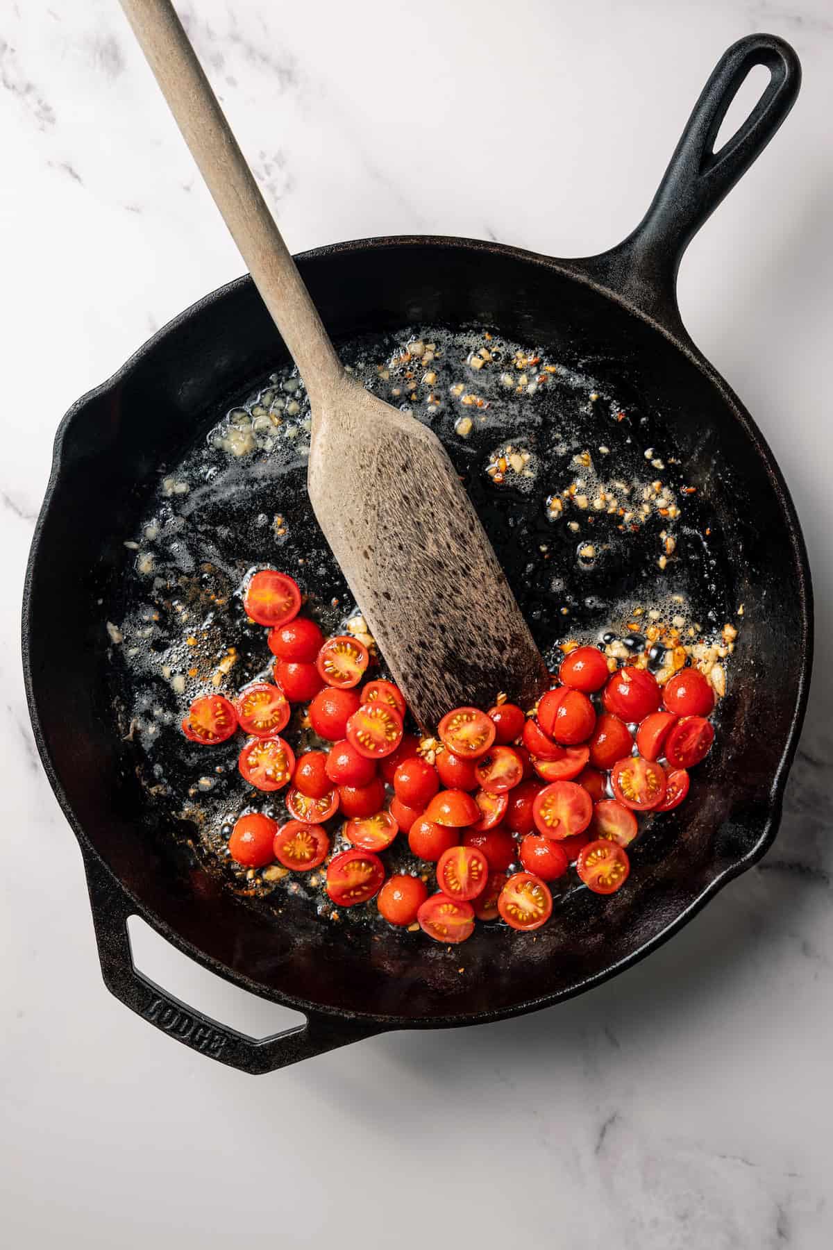 A wooden spoon stirring sautéed cherry tomatoes in a skillet.