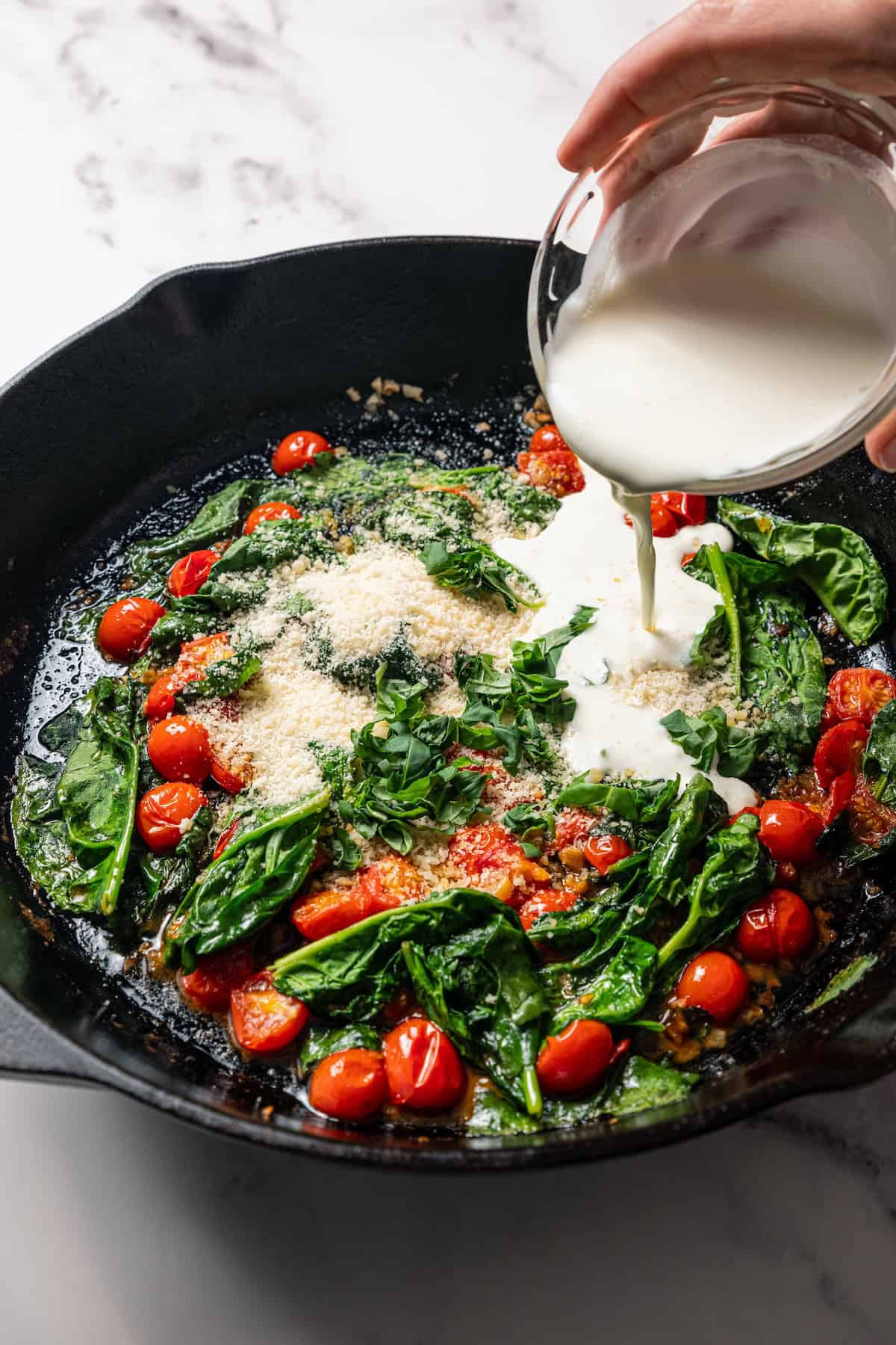 Heavy cream being poured over sautéed spinach and tomatoes in a skillet.
