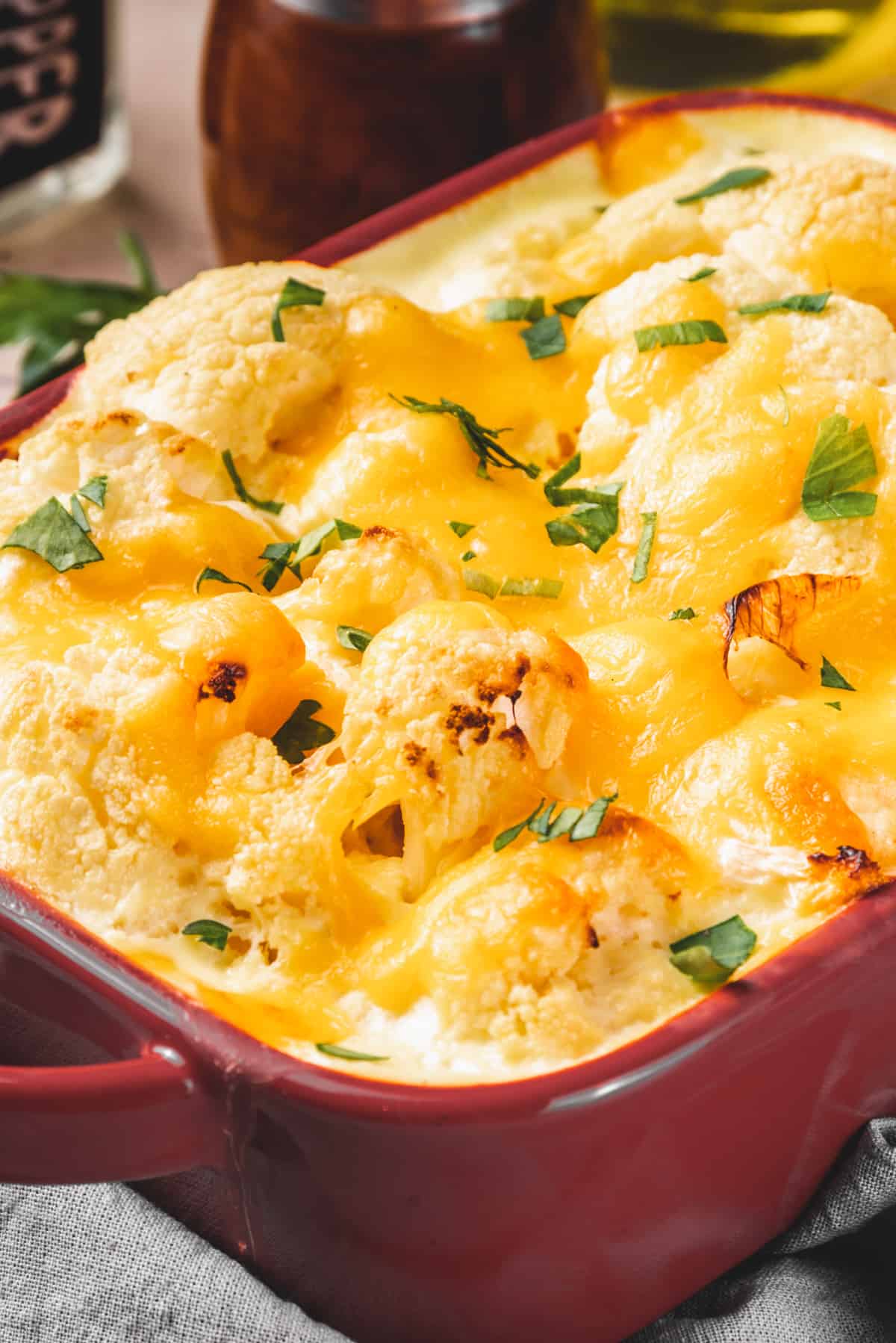 Side shot of a dark red baking dish with a loaded cheesy cauliflower casserole in creamy sauce.
