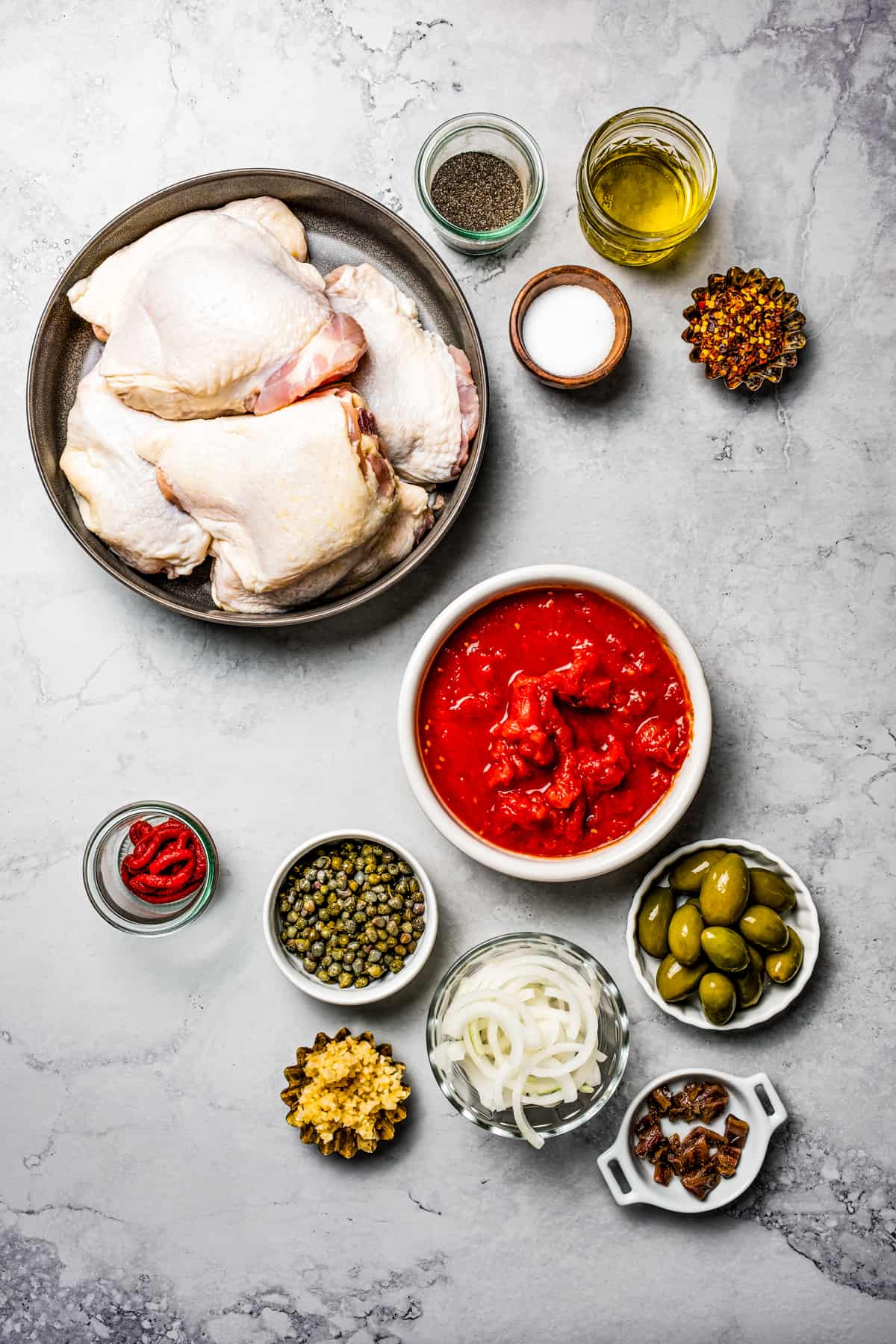 From top: Chicken thighs, seasonings, olive oil, anchovies, capers, tomatoes, olives, garlic, sliced onion, spices.