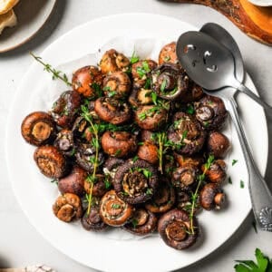 Overhead view of roasted mushrooms garnished with fresh thyme sprigs on a white plate with two serving spoons.