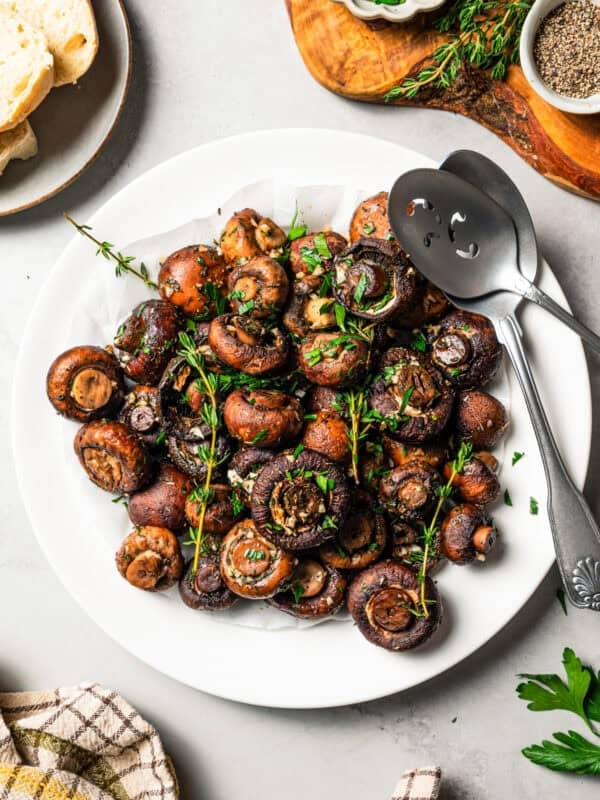 Overhead view of roasted mushrooms garnished with fresh thyme sprigs on a white plate with two serving spoons.