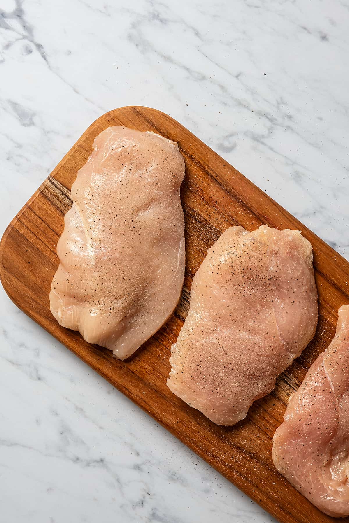 A cutting board with three chicken cutlets arranged on it, sprinkled with salt and pepper.