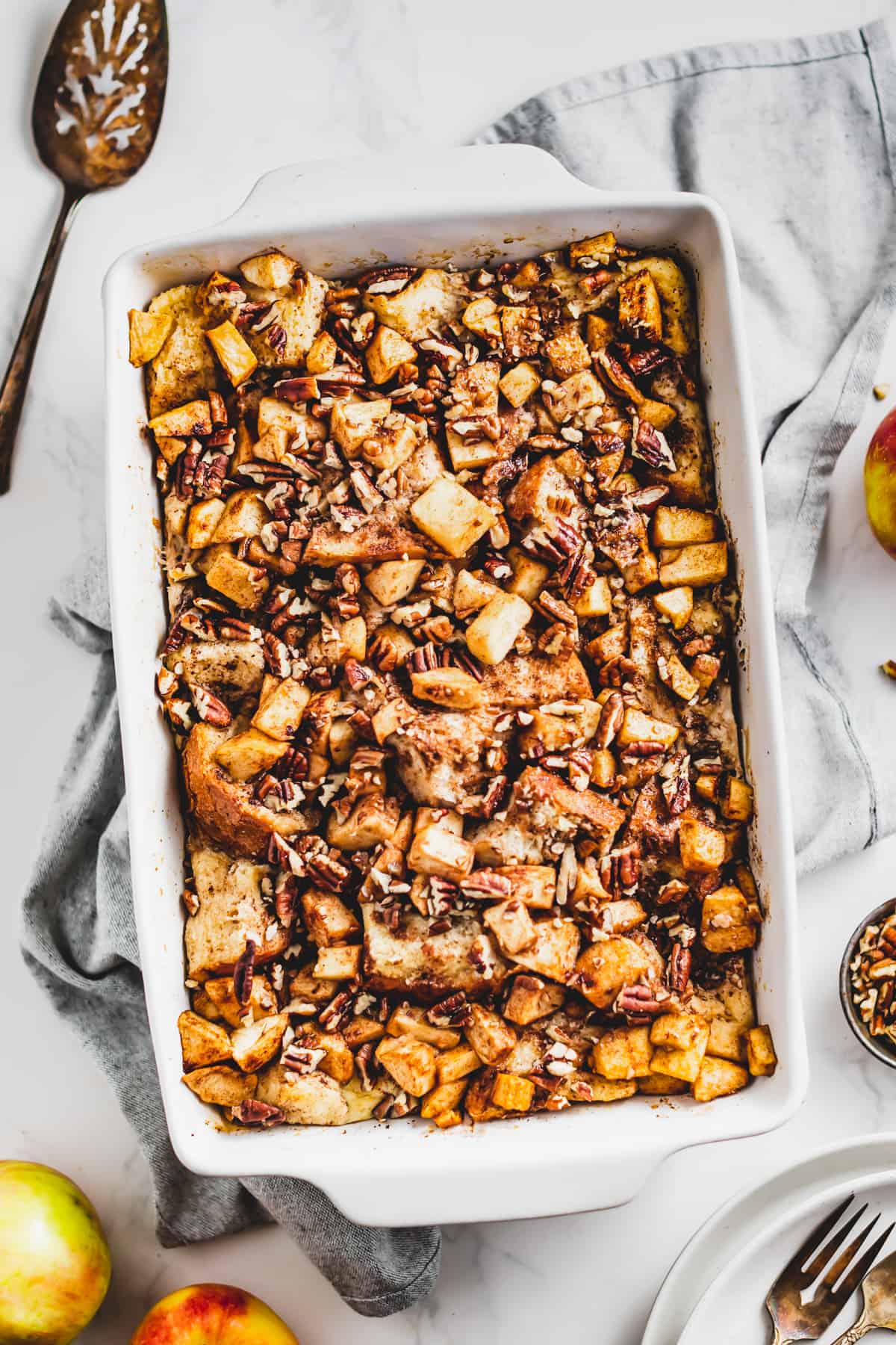 overhead wide shot of Baked apple cinnamon French toast in a baking dish, with a large spoon placed on the top left and fresh apples on the bottom left of the baking dish.