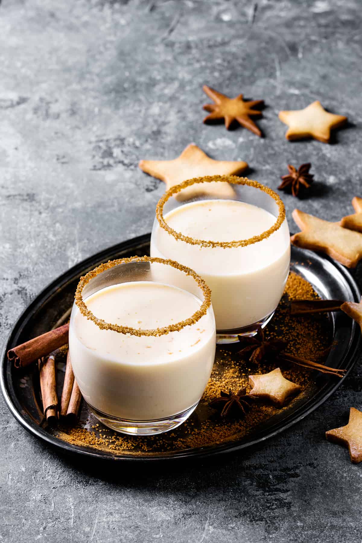 Eggnog cocktail with cinnamon, served in two glasses with star shape sugar cookies, fir branch over gray background.