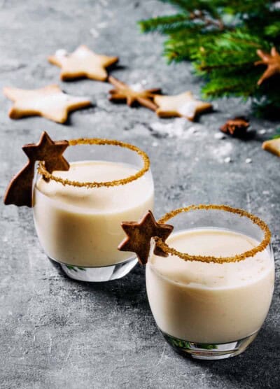 Eggnog cocktail with cinnamon, served in two glasses with star shape sugar cookies, fir branch over gray background.