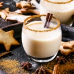 Eggnog Christmas milk cocktail with cinnamon, served in two glasses with shortbread star shape sugar cookies different size, decor toys, fir branch over dark texture background. Close up