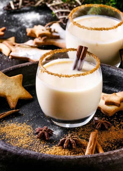 Eggnog Christmas milk cocktail with cinnamon, served in two glasses with shortbread star shape sugar cookies different size, decor toys, fir branch over dark texture background. Close up