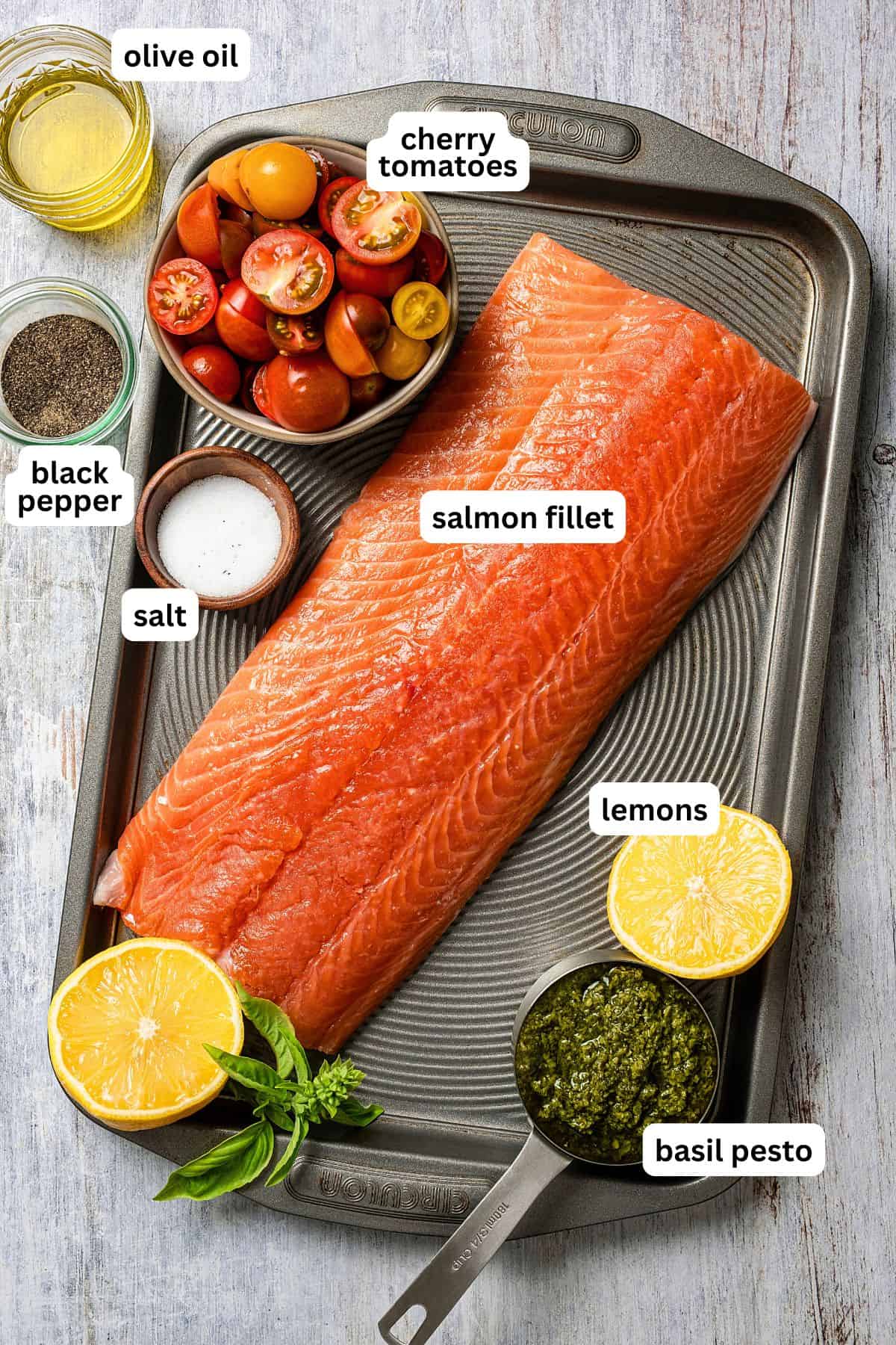 Raw salmon filet on a baking sheet surrounded by ingredients to make pesto salmon including cherry tomatoes, basil pesto, salt, pepper, oil, and lemons.