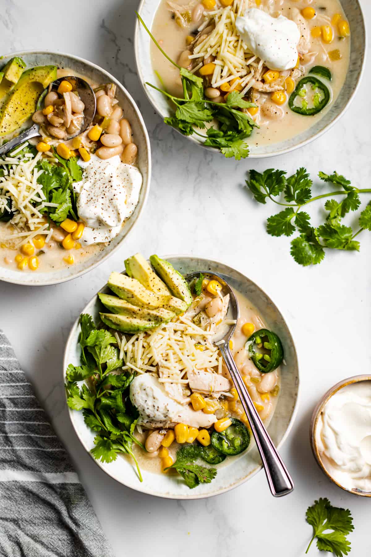 overhead shot of three soup bowls with homemade turkey chili and garnished with jalapeno slices, shredded parmesan, avocado slices, corn kernels, and sour cream.