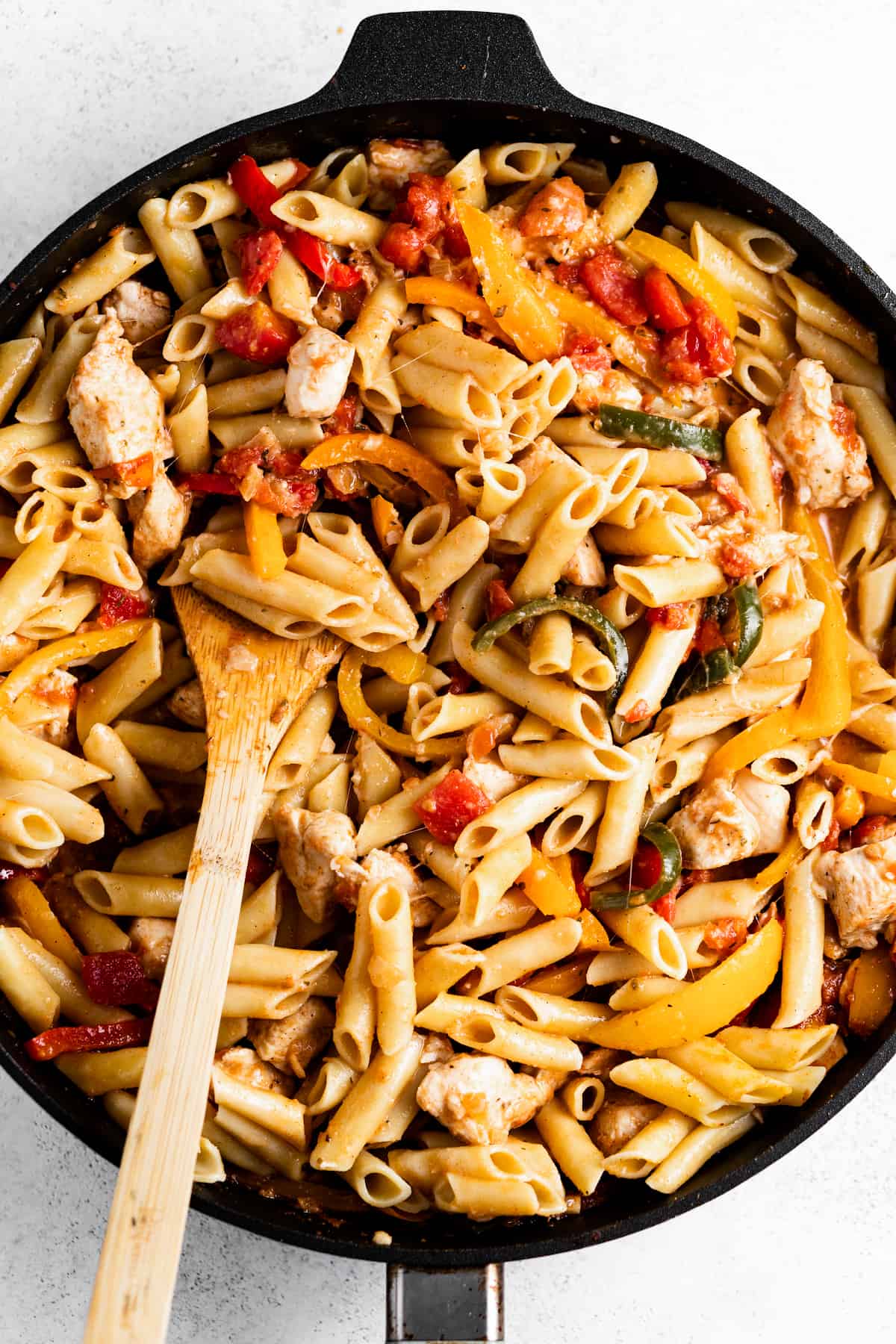 a skillet with pasta, chicken, and sliced peppers, with a wooden spoon stirring through.