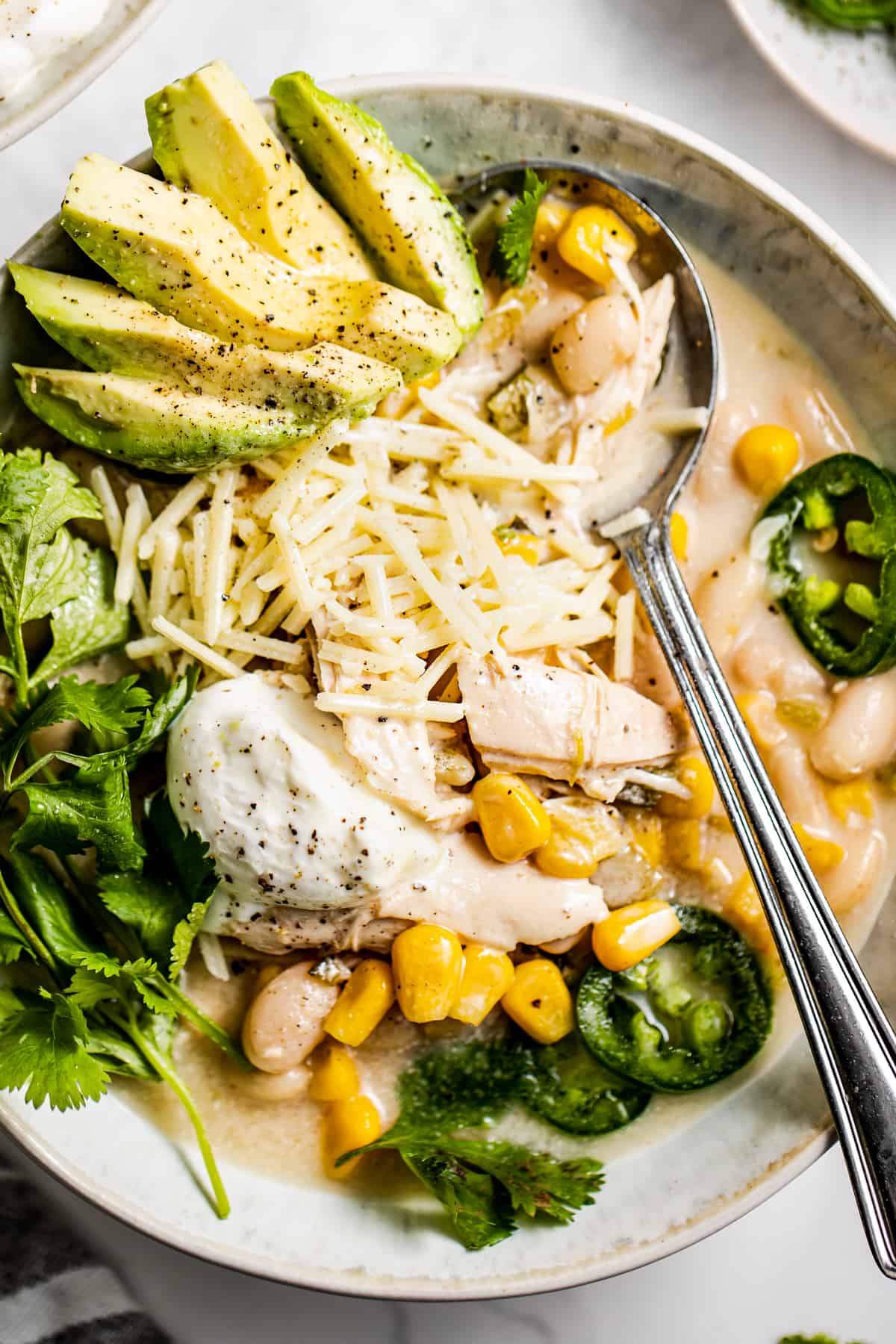 Up close shot of a soup bowl with homemade turkey chili and garnished with jalapeno slices, shredded parmesan, avocado slices, corn kernels, and sour cream.