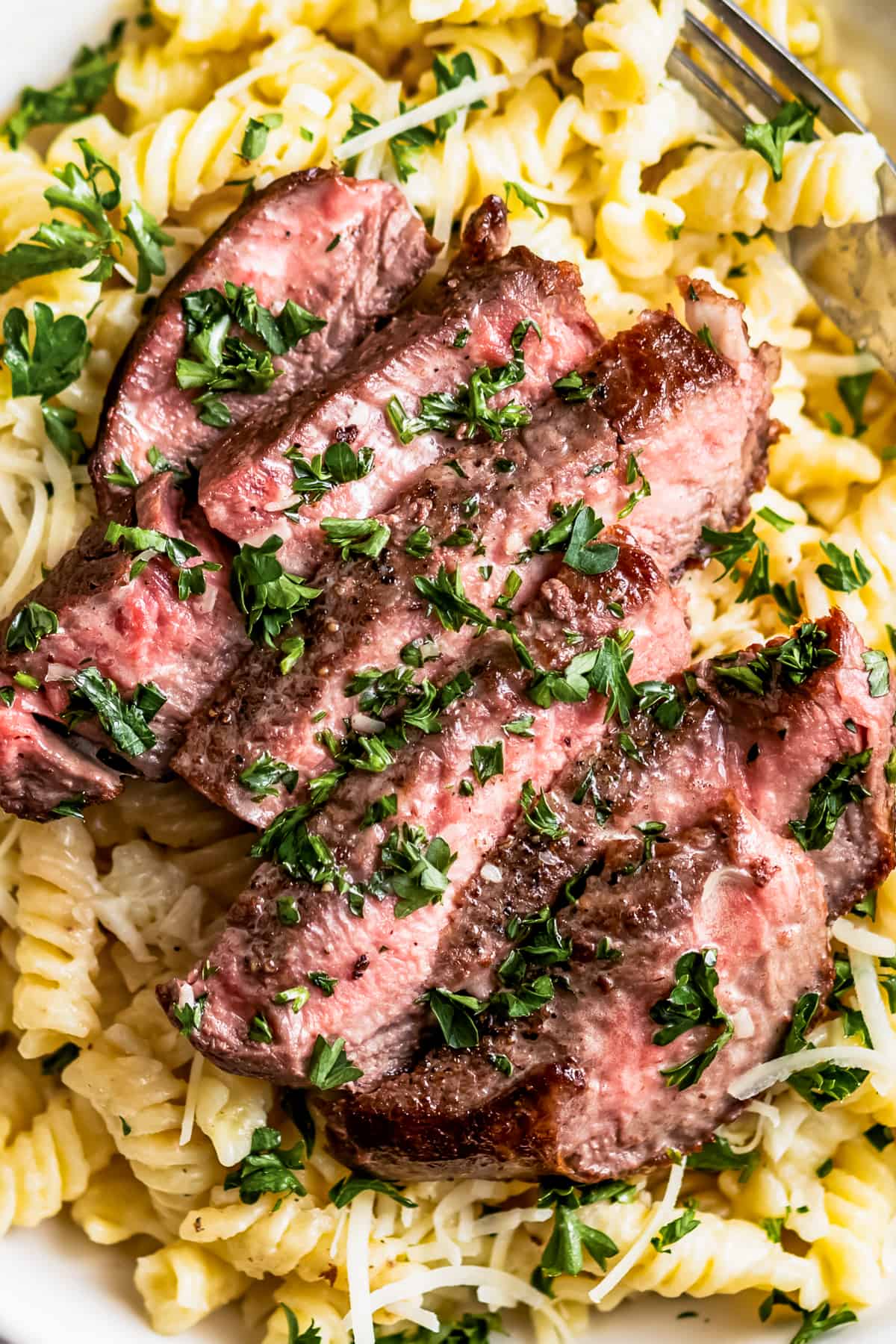 Overhead shot of a sliced steak set atop Alfredo pasta with parmesan and parsley.