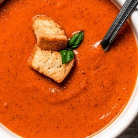 close up shot of a bowl of creamy tomato bisque garnished with croutons and basil leaves, a spoon stirring through it.