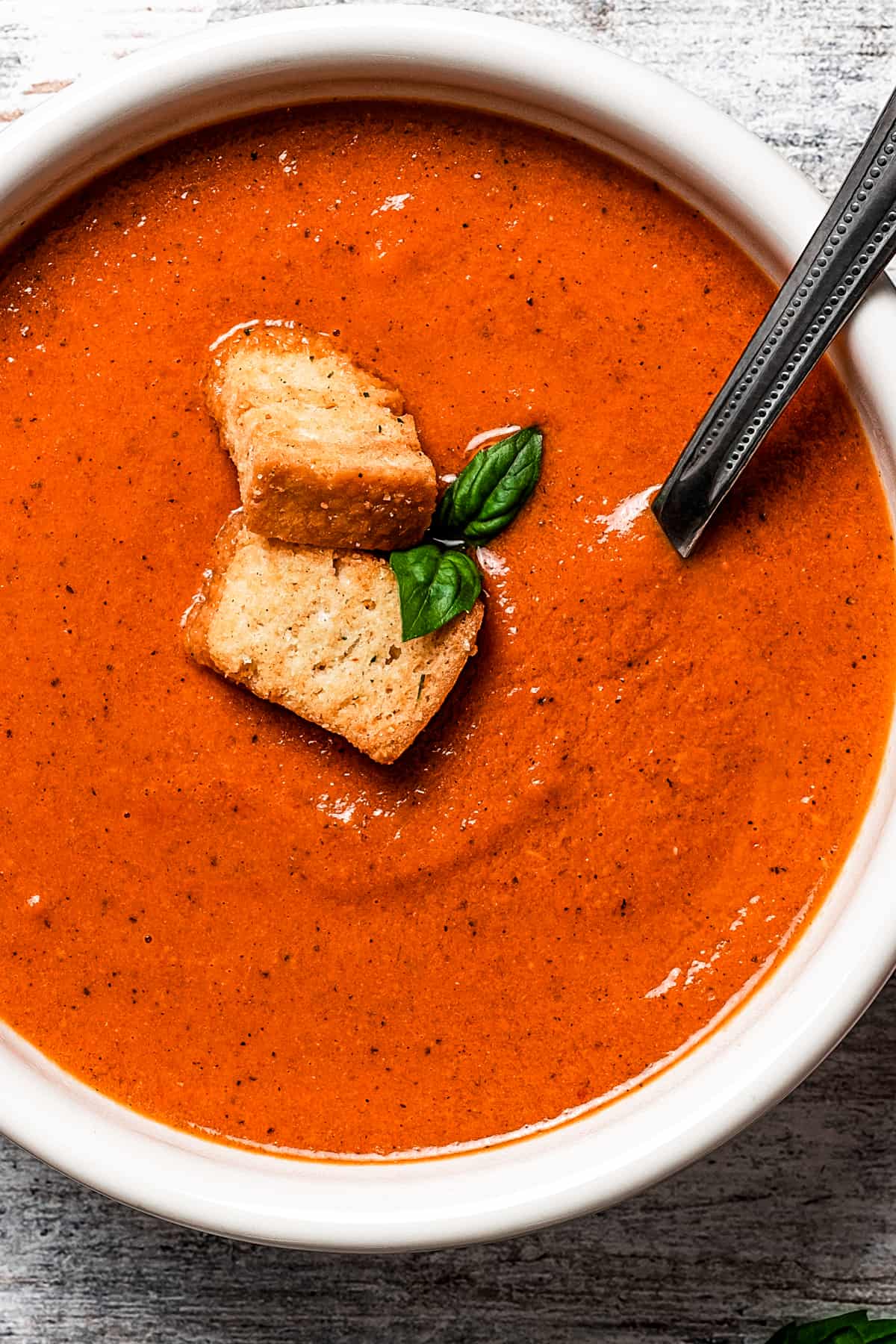 close up shot of a bowl of creamy tomato bisque garnished with croutons and basil leaves, a spoon stirring through it.