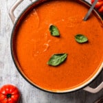 overhead shot of a soup pot with tomato bisque, topped with three basil leaves, and tomatoes and croutons placed around the pot.