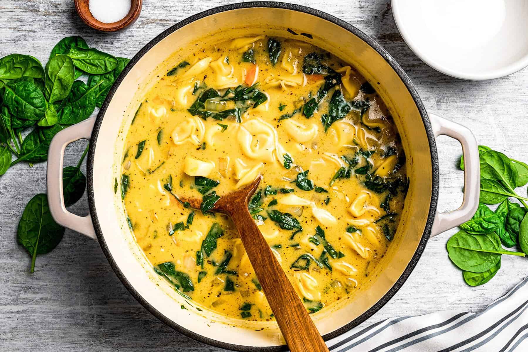This creamy Tortellini Soup with spinach is an easy vegetarian soup recipe packed with tender cheese tortellini, fresh veggies, and lots of flavor! A pot of creamy tortellini soup, with a wooden spoon stirring through it.