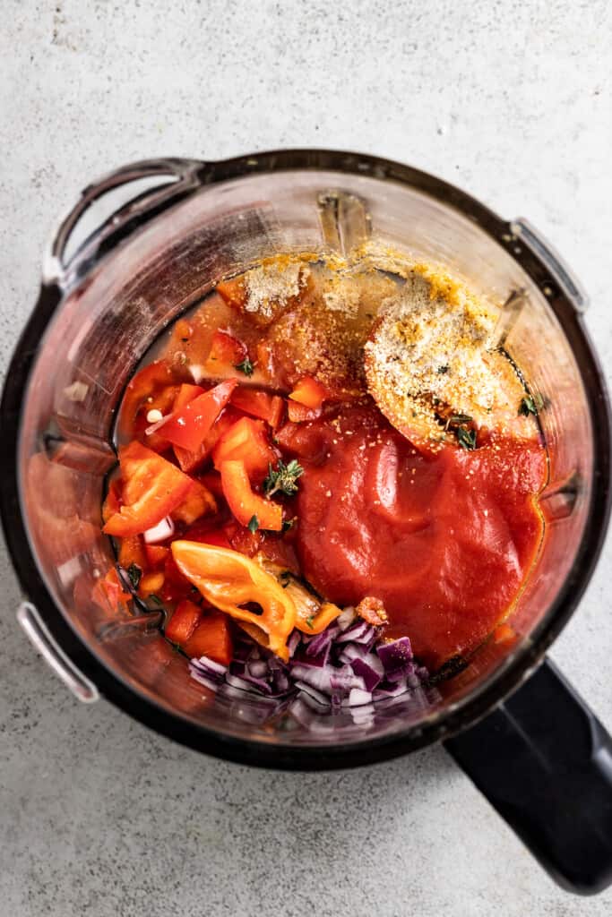 Red onion, red bell pepper, tomato puree, Roma tomato, garlic powder, thyme, Scotch bonnet, chicken bouillon granules, chicken broth, and Nigerian curry powder in a blender ready to be blended.