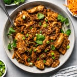 A bowl with chicken bulgogi and smaller bowls arranged around the bowl with carrot sticks, fresh cilantro leaves, and white rice.
