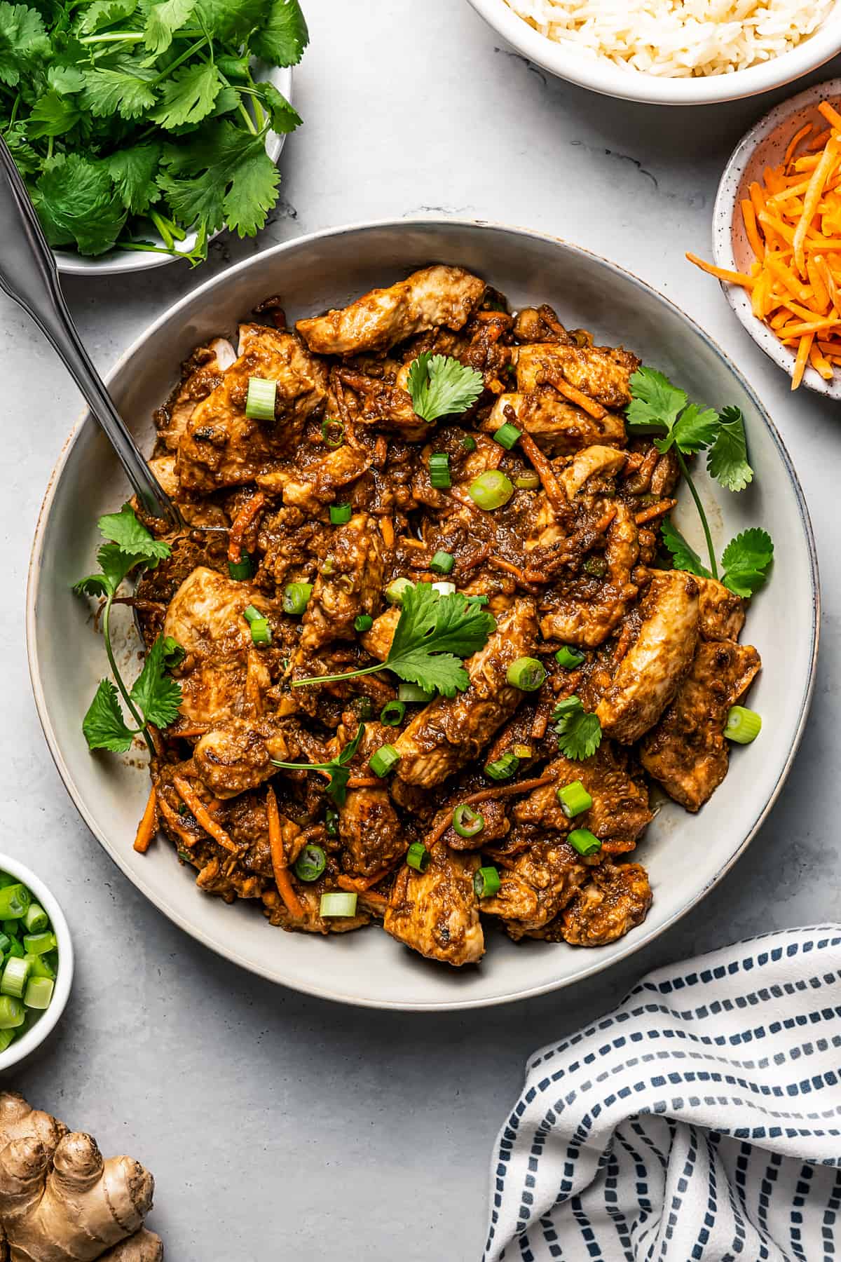 A bowl with chicken bulgogi and smaller bowls arranged around the bowl with carrot sticks, fresh cilantro leaves, and white rice.