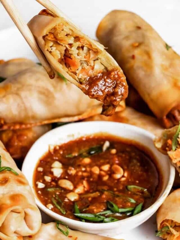 chopsticks holding up a spring roll over a bowl of peanut sauce.