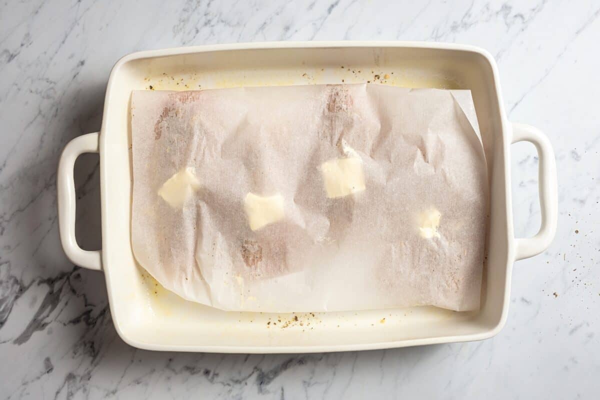 Four chicken breasts are placed in a baking dish and covered with a sheet of parchment paper.