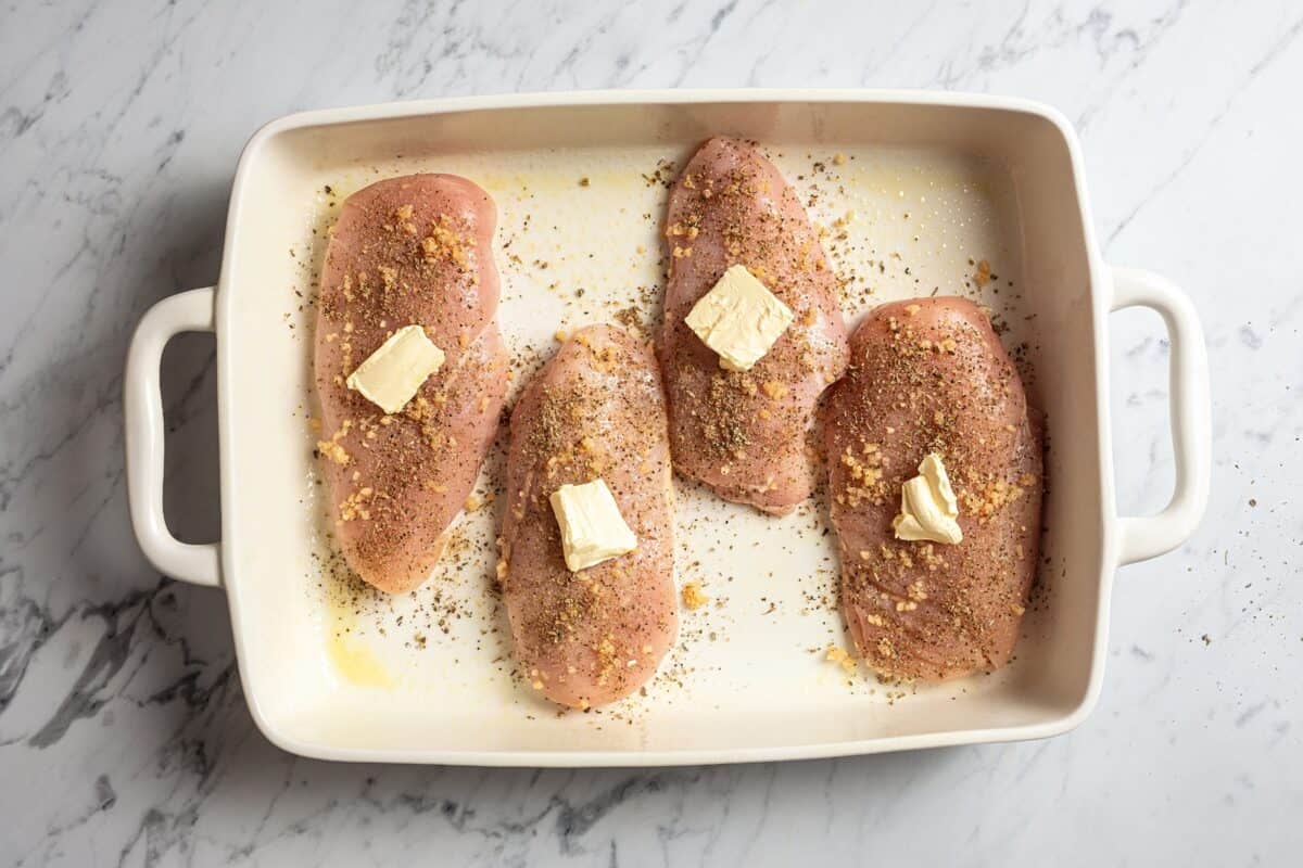 Four chicken breasts in a baking dish, each topped with a pat of butter.