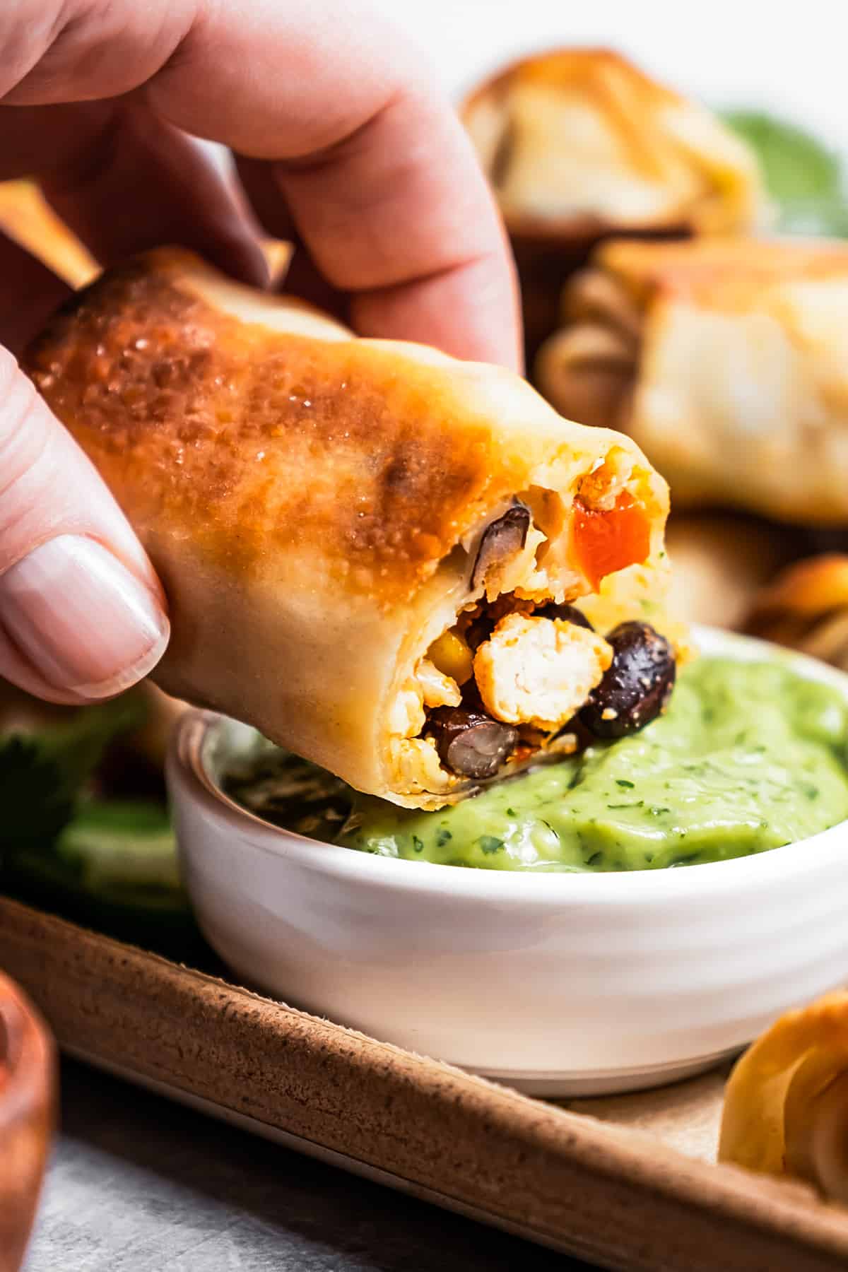 Close-up of a hand dipping homemade egg rolls in avocado crema.