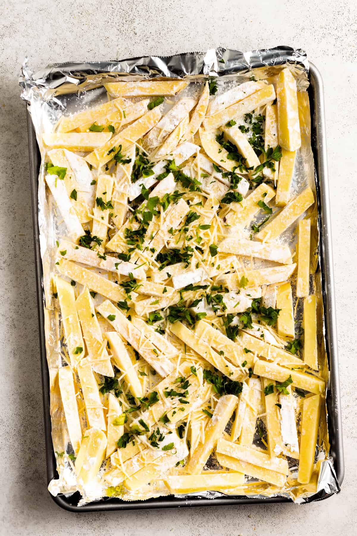 Potatoes are cut into fries, topped with fresh chopped parsley, and arranged on a baking sheet.