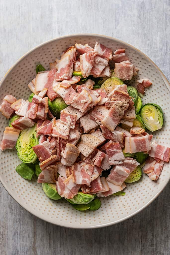 Adding diced bacon to halved Brussels sprouts in a mixing bowl.