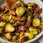 Scooping roasted Brussels sprouts and bacon out of a serving bowl with a wooden serving fork.