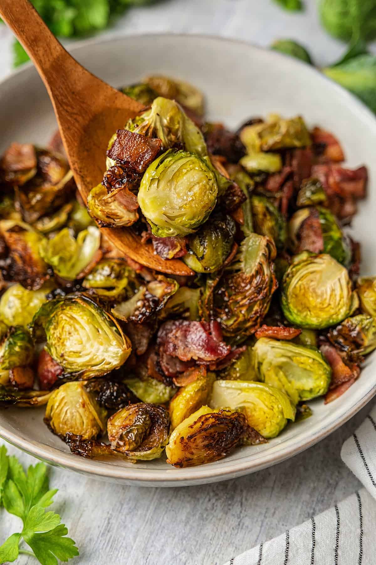 Scooping roasted Brussels sprouts and bacon out of a serving bowl with a wooden serving fork.