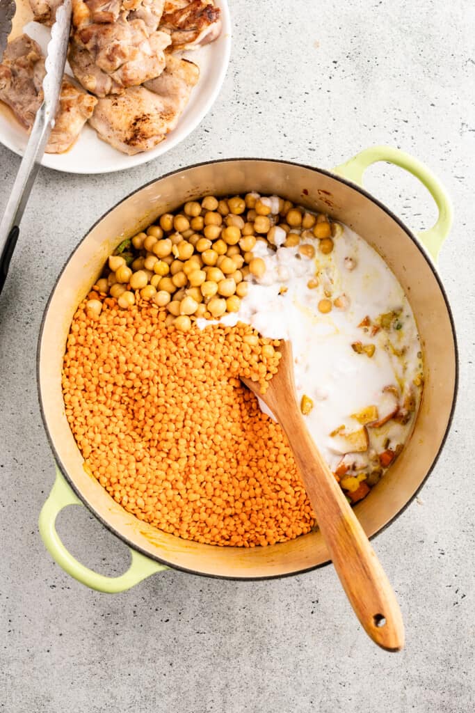 Adding lentils, coconut milk, and chickpeas to a stew pot near a bowl of browned chicken.