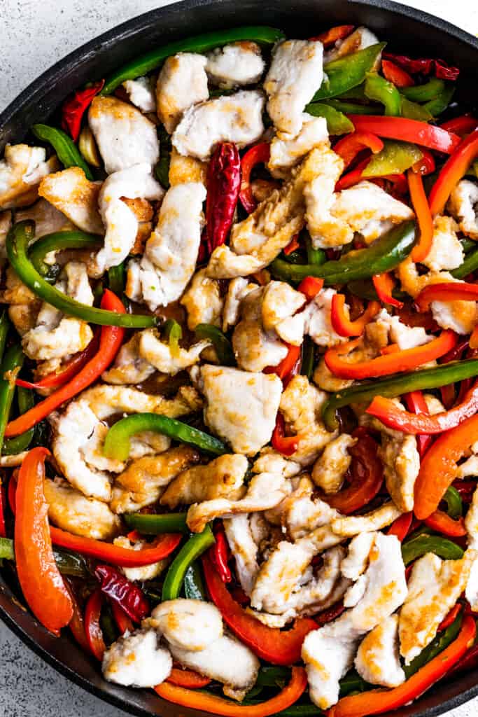 Browned chicken, sautéed bell peppers, scallions, and dried red chilis in a pan.