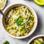 Two bowls of pozole verde in bowls with garnishes near cilantro, avocado, and shredded cabbage.