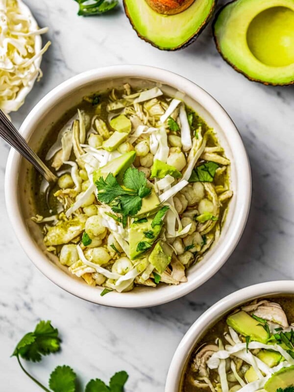 Two bowls of pozole verde in bowls with garnishes near cilantro, avocado, and shredded cabbage.