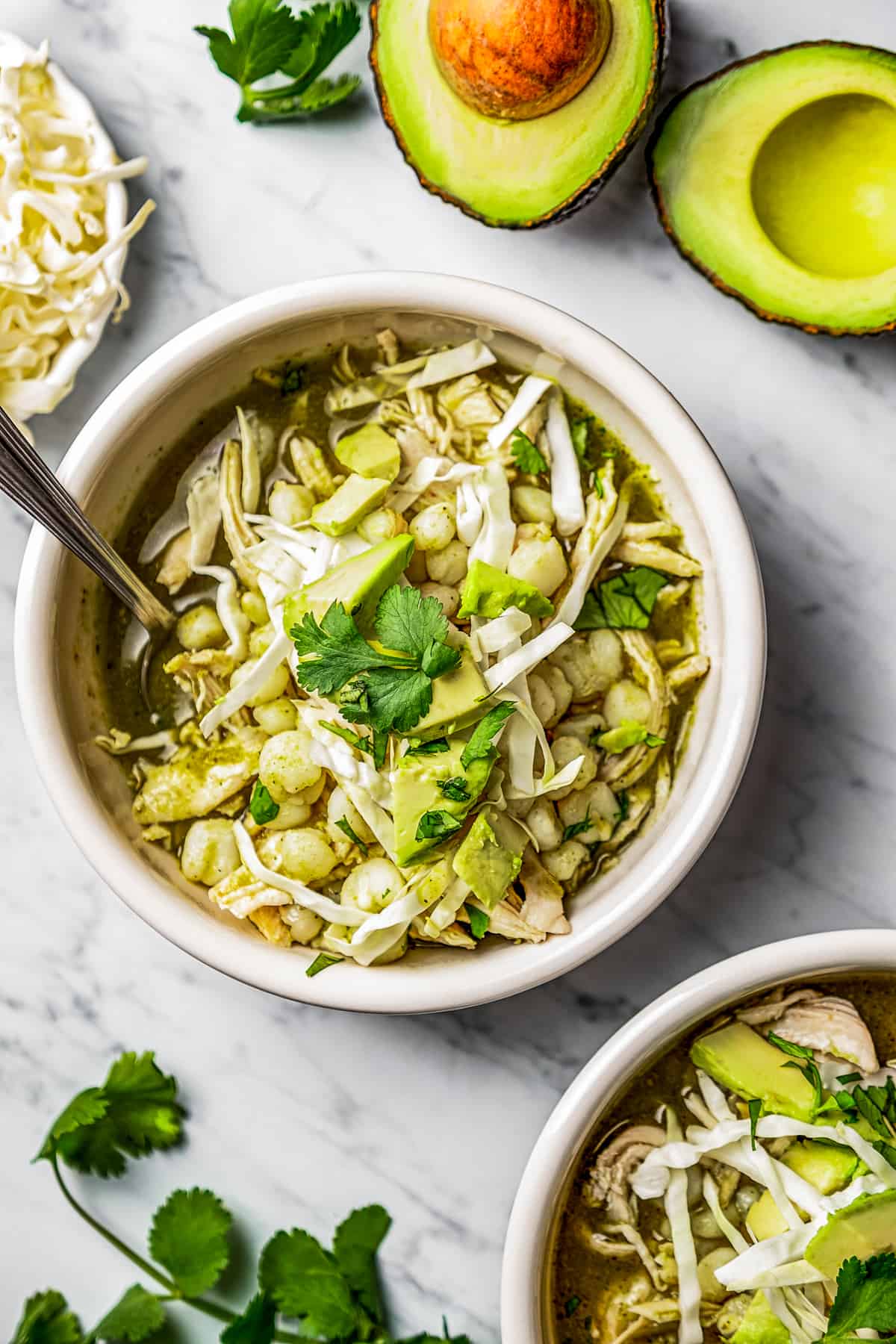 Two bowls of pozole verde in bowls with garnishes near cilantro, avocado, and shredded cabbage.