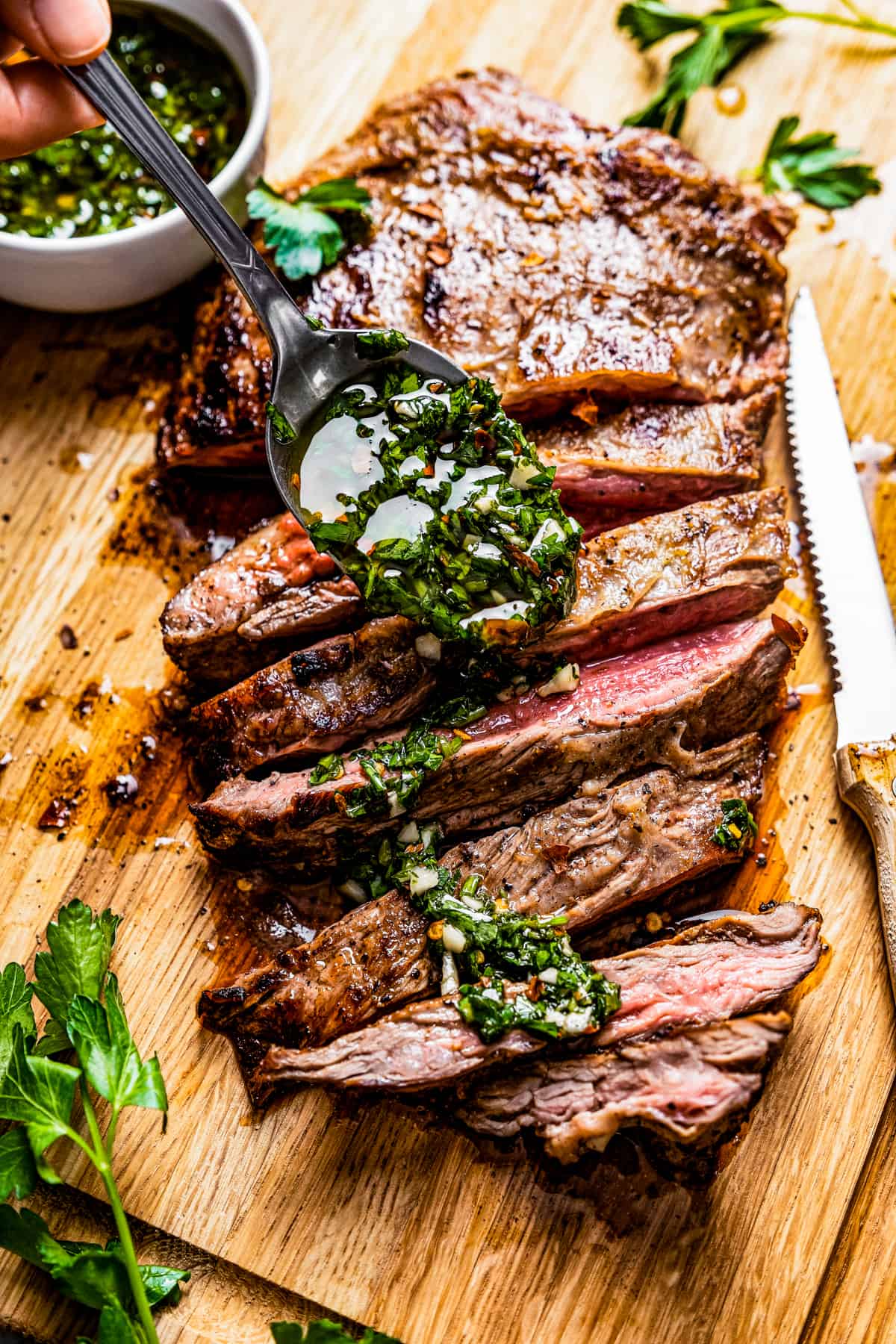 Spooning chimichurri over flank steak on a cutting board.