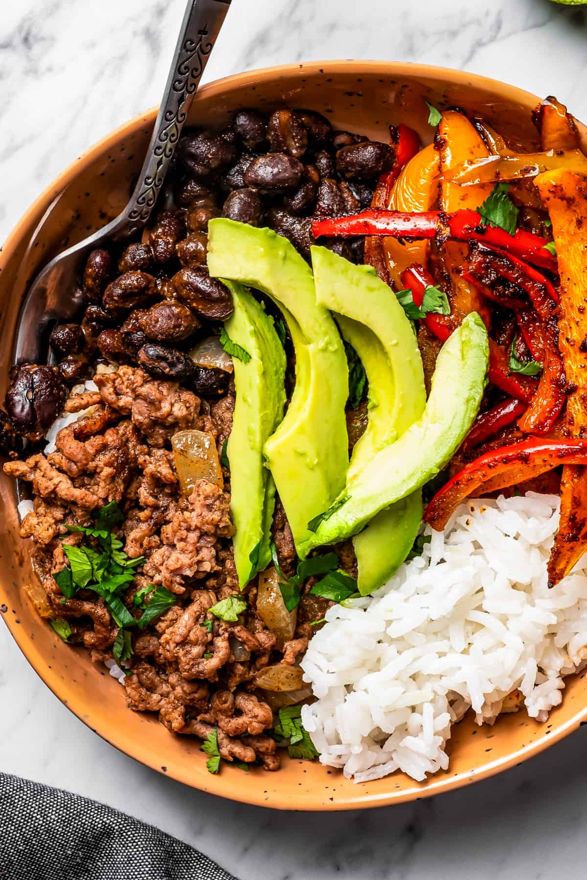Closeup of a bowl with taco meat, sliced bell peppers, avocado slices, beans, and rice.