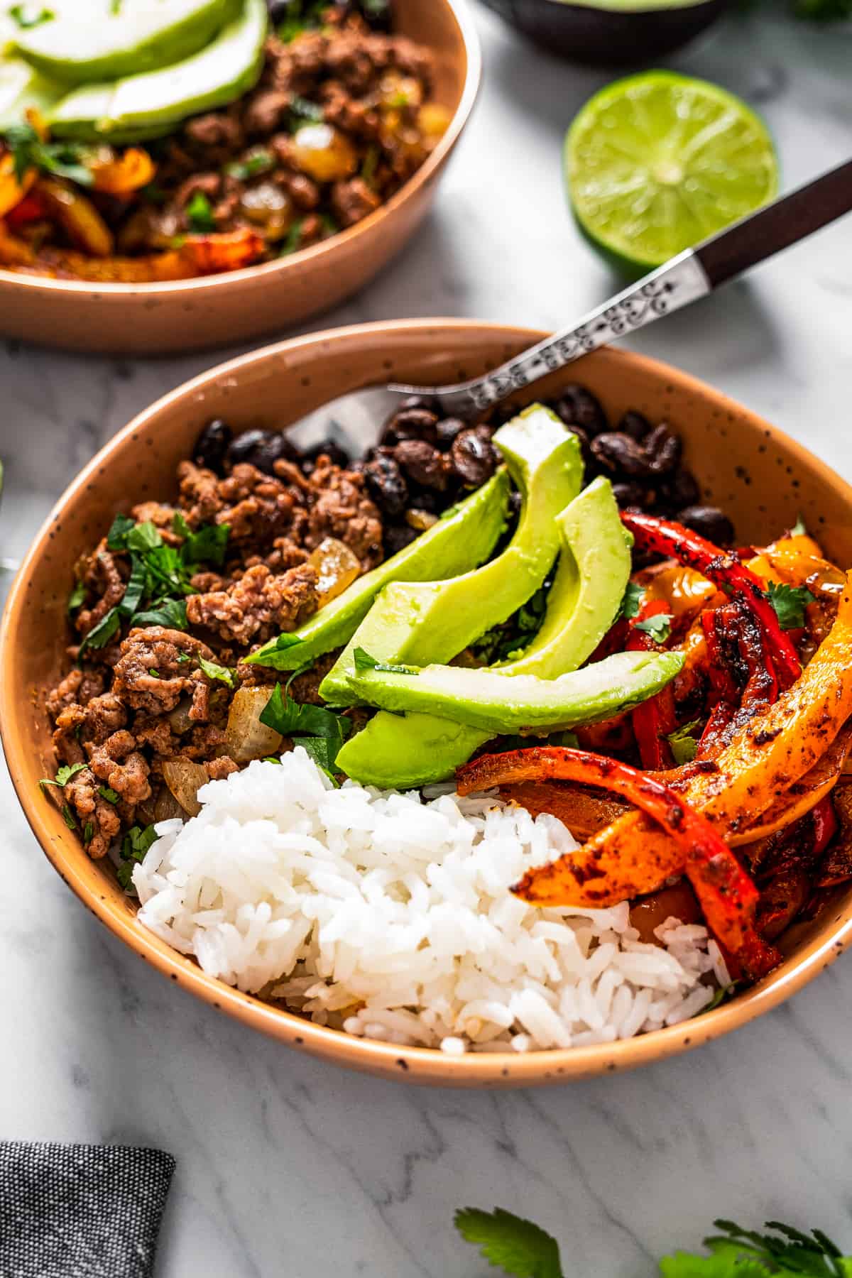 Two taco bowls with taco meat, sliced bell peppers, avocado slices, beans, and rice.