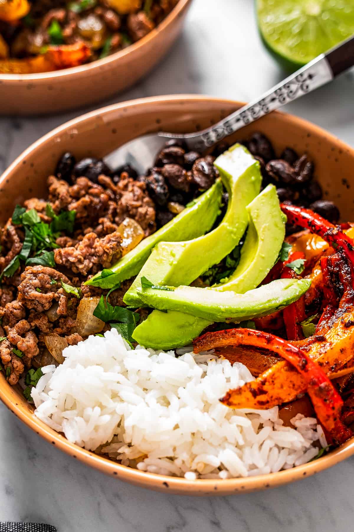 side shot of a bowl with taco meat, sliced bell peppers, avocado slices, beans, and rice.