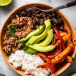 overhead shot of a taco bowl with taco meat, sliced bell peppers, avocado slices, beans, and rice, and a fork placed inside the bowl.