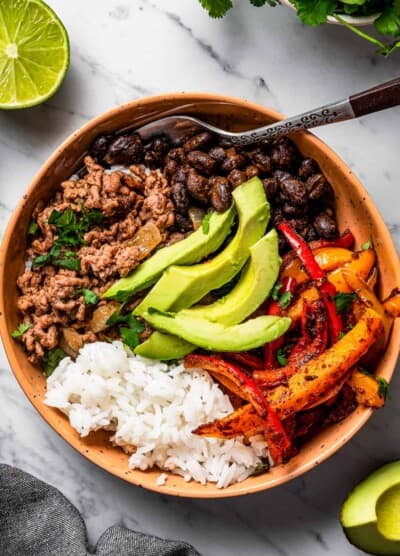 overhead shot of a taco bowl with taco meat, sliced bell peppers, avocado slices, beans, and rice, and a fork placed inside the bowl.