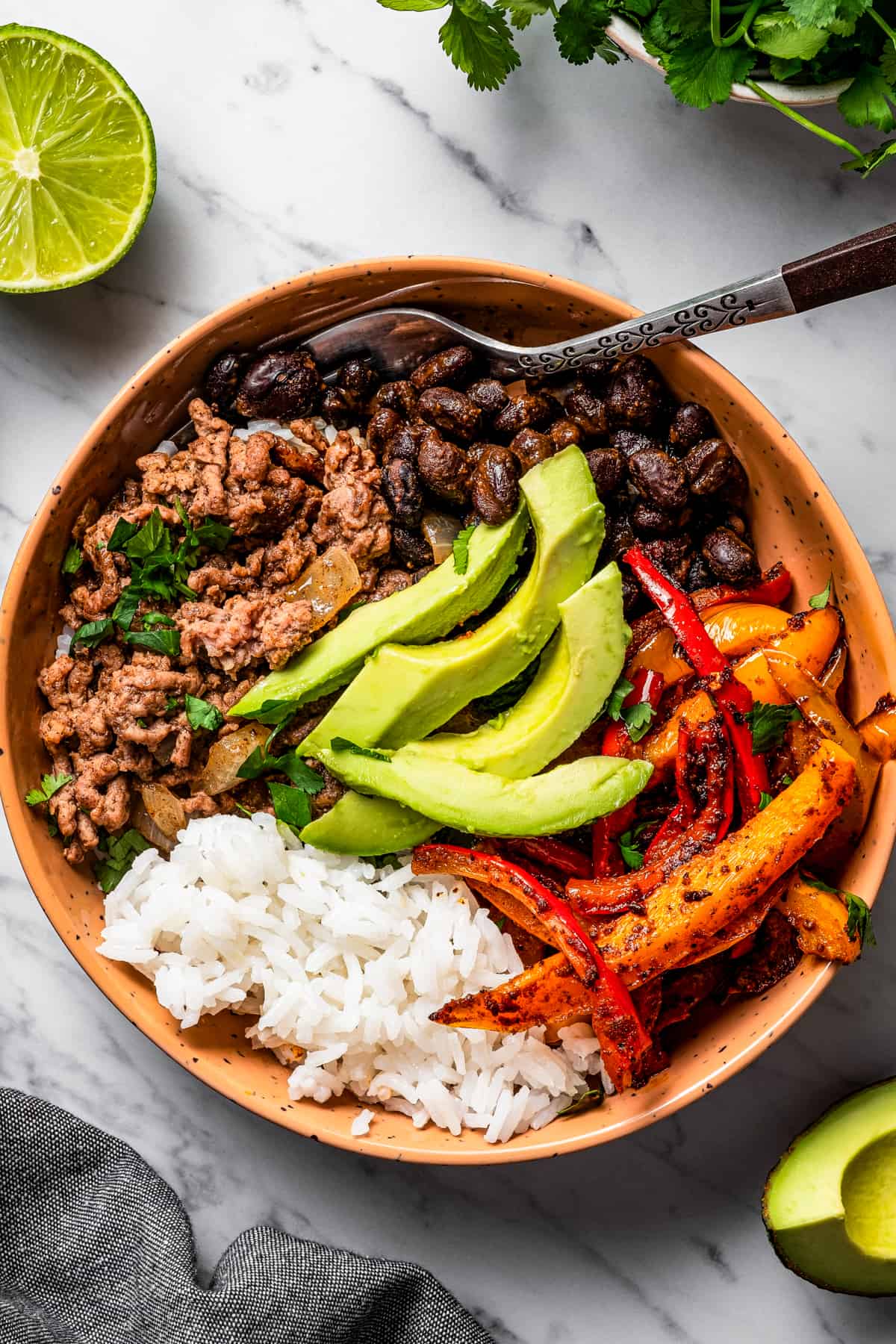 overhead shot of a taco bowl with taco meat, sliced bell peppers, avocado slices, beans, and rice, and a fork placed inside the bowl.