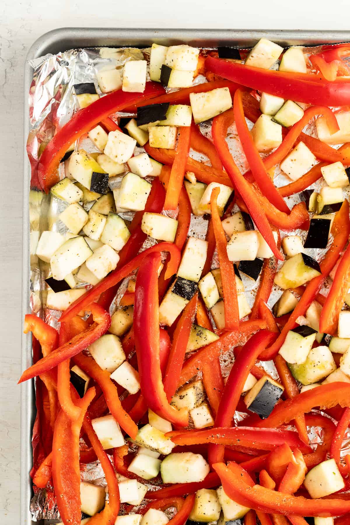 Raw cubed eggplant and sliced red bell pepper on a baking sheet lined with aluminum foil.