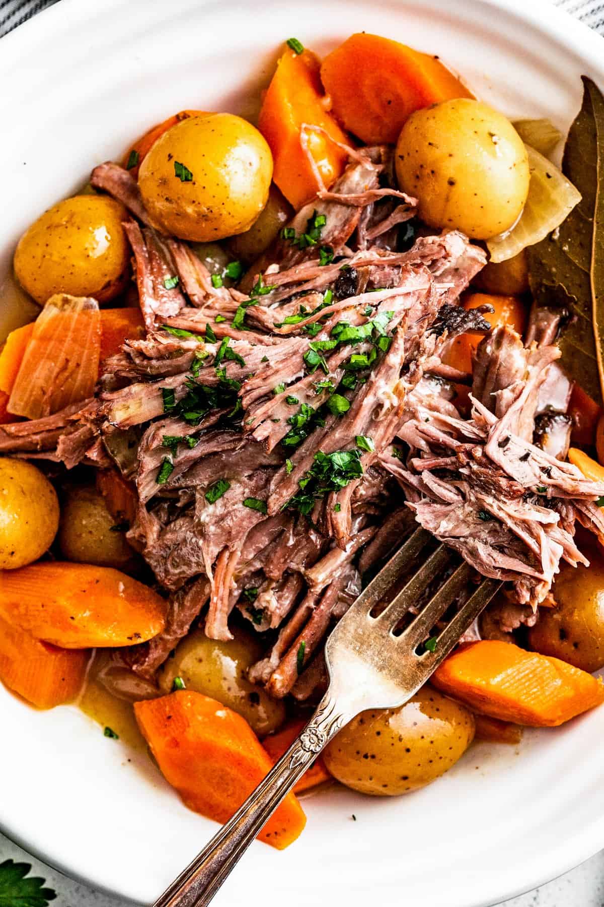 Shredded rump roast served on a plate with potatoes and carrots, and a fork.