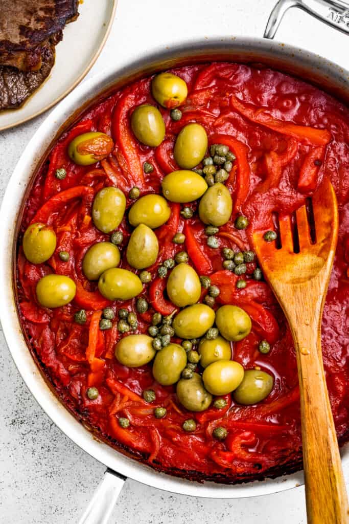 Adding olives and capers to tomato sauce in a pan.