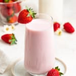 Strawberry milk in a glass with a strawberry on the edge of the glass. Fresh strawberries and milk are behind it.